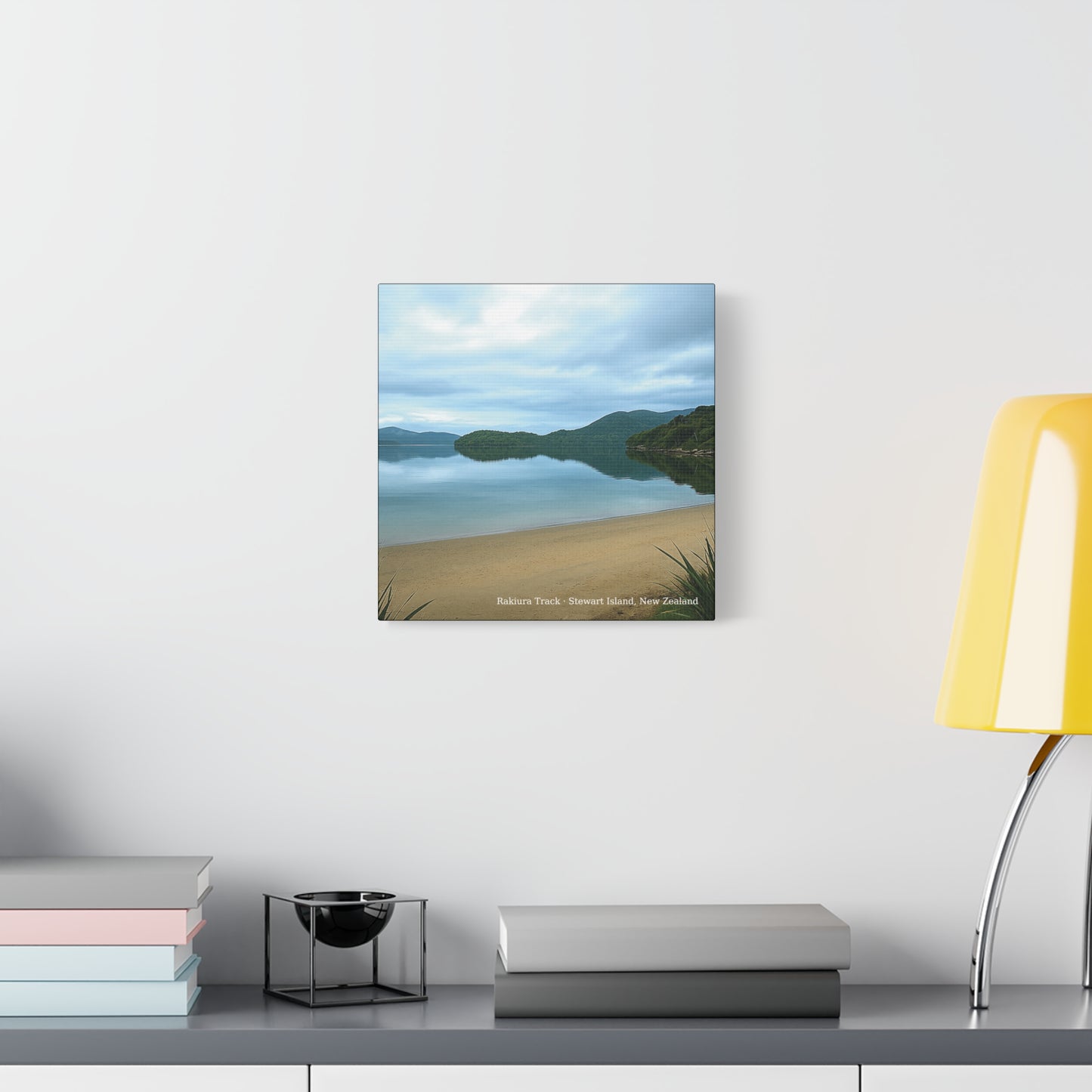 Framed landscape print of a beach and mountain scene on a wall above a desk with books and a lamp.