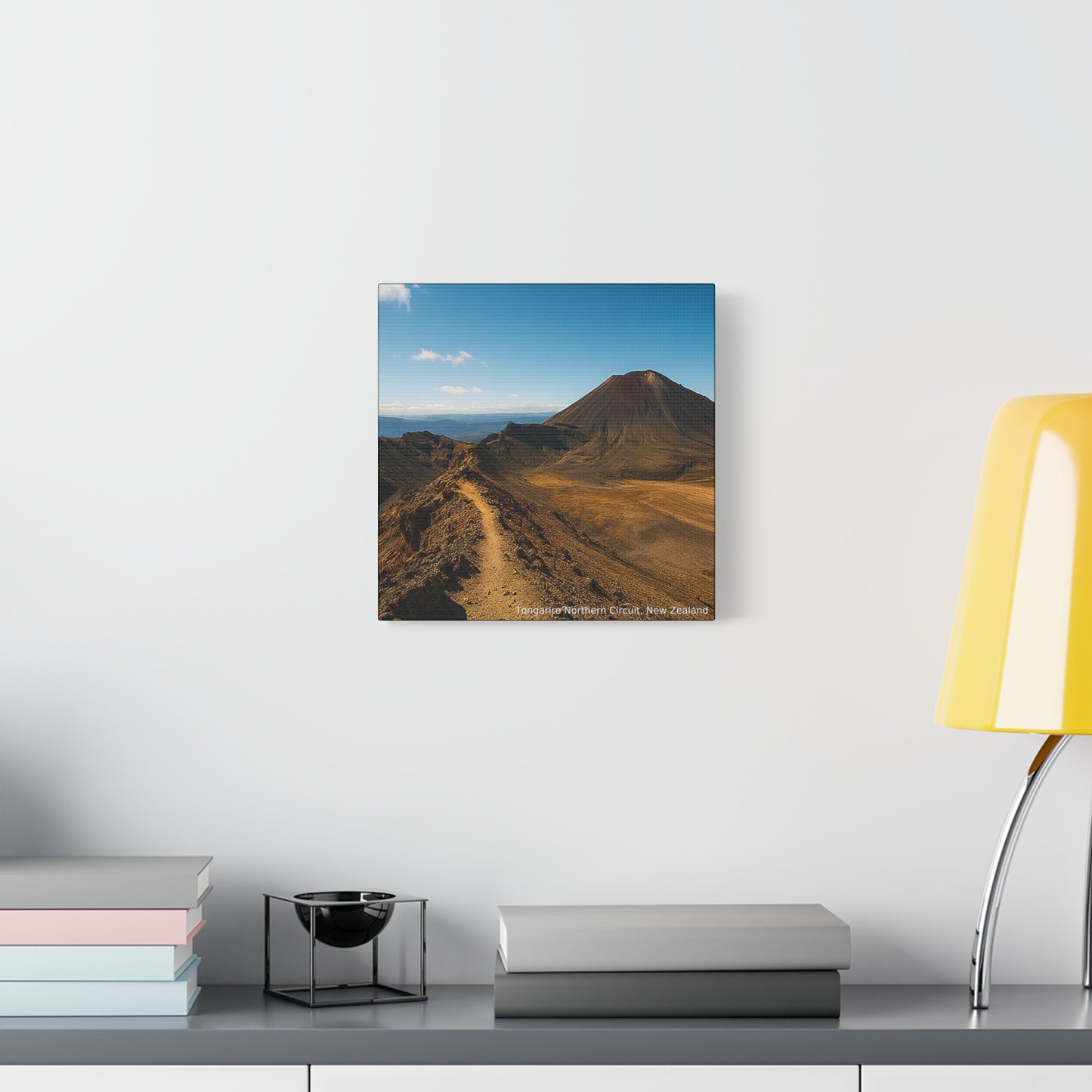 Tongariro Northern Circuit Mountains landcape painting on a wall above a shelf with books and a lamp.