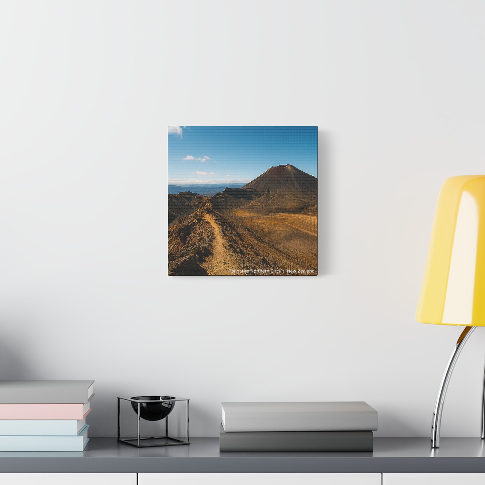 Tongariro Northern Circuit Mountains landcape painting on a wall above a shelf with books and a lamp.