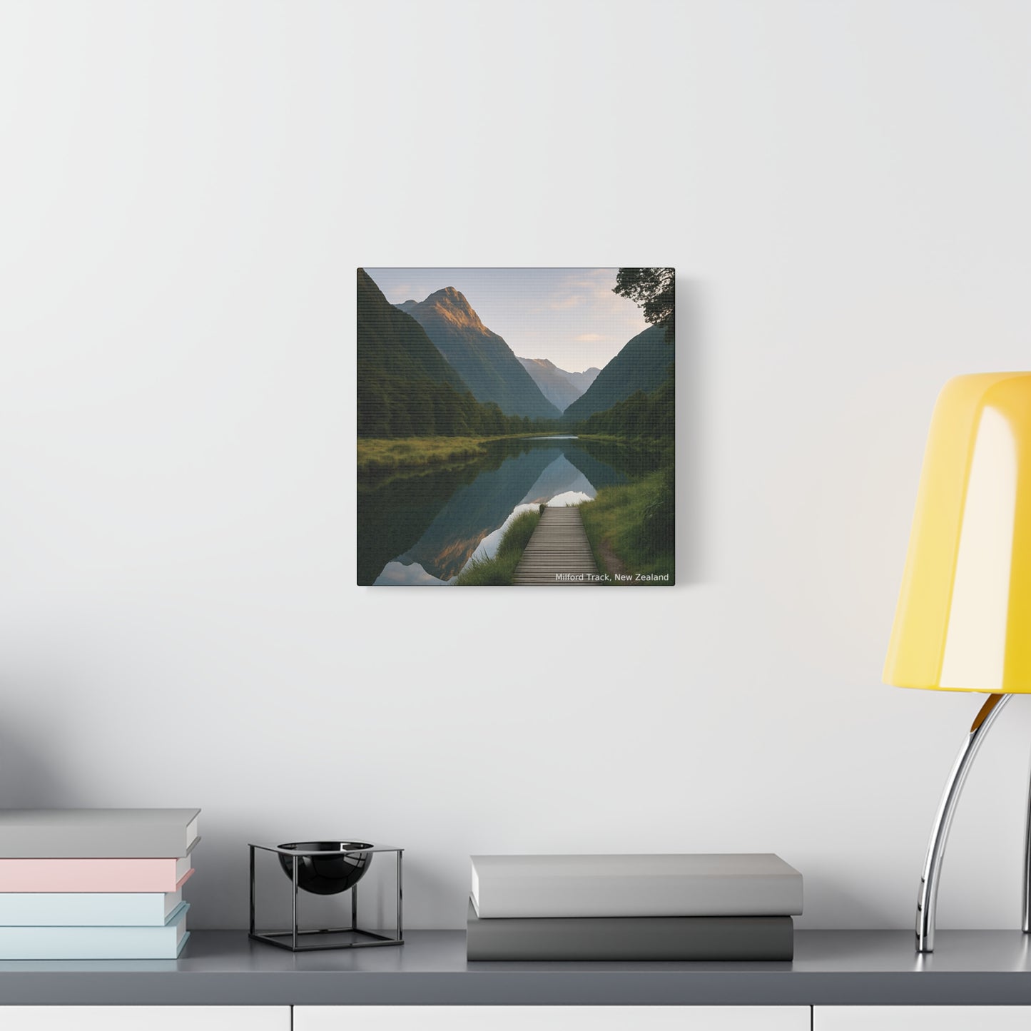 Framed Milford Track landscape print of a mountainous valley with a lake on a white wall above a desk.