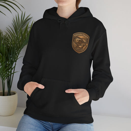 Person wearing a black Rakiura Track hoodie with a gold emblem in a minimal indoor setting