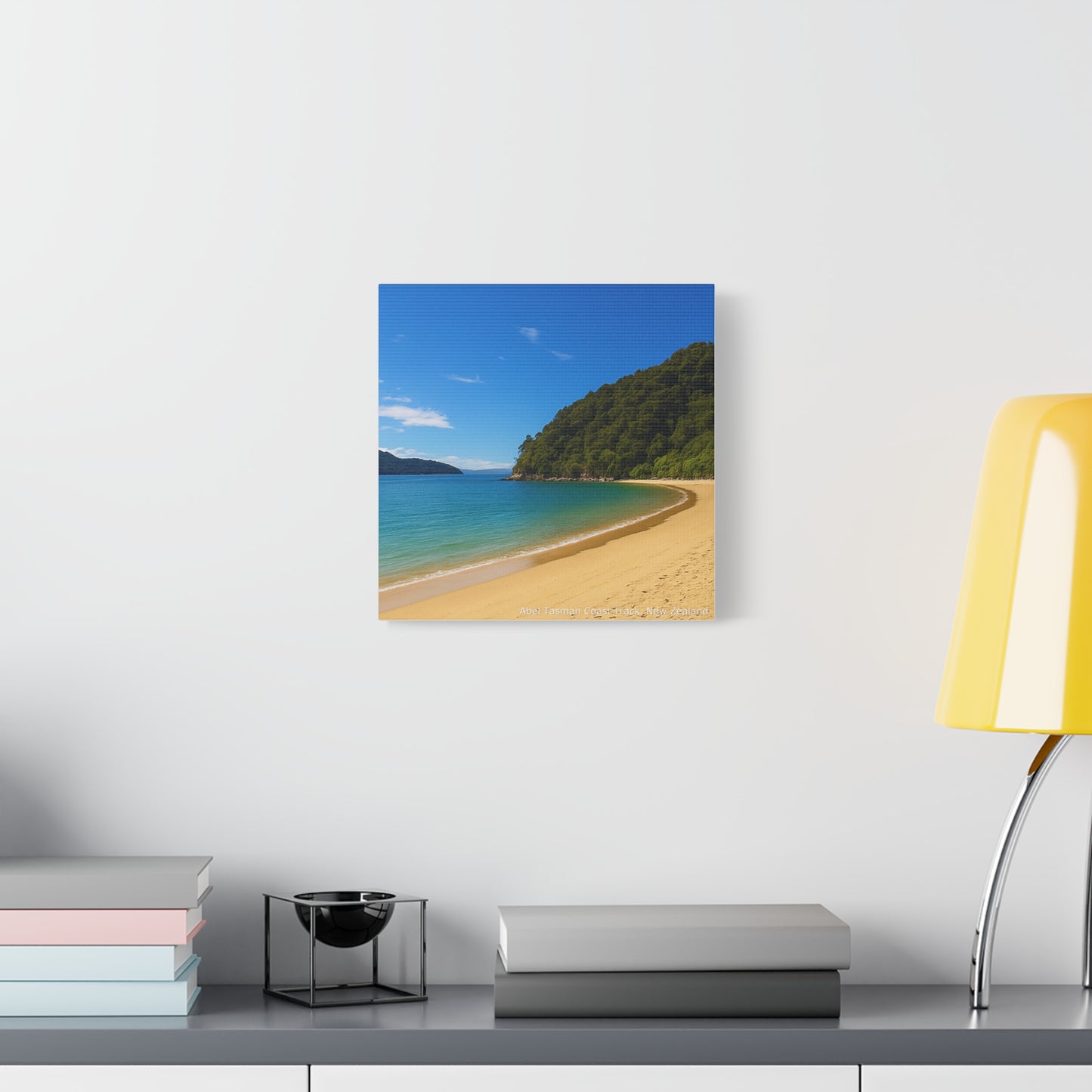 Abel Tasman Beach scene wall art on a white wall with books and a lamp.