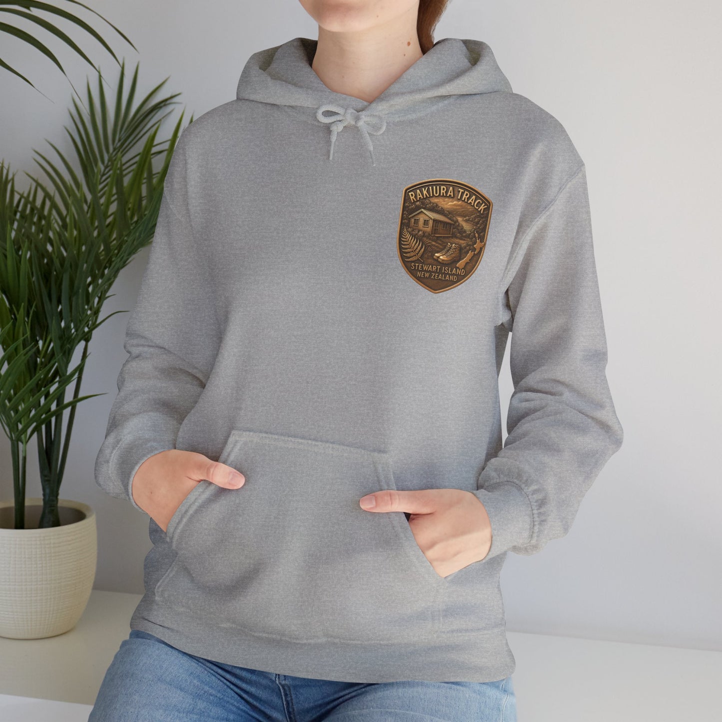 Person wearing a gray Rakiura Track hoodie with a logo in a room with a plant