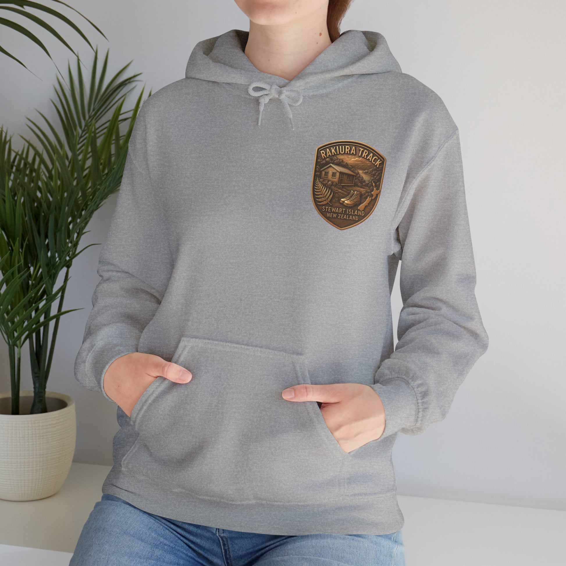 Person wearing a gray Rakiura Track hoodie with a logo in a room with a plant