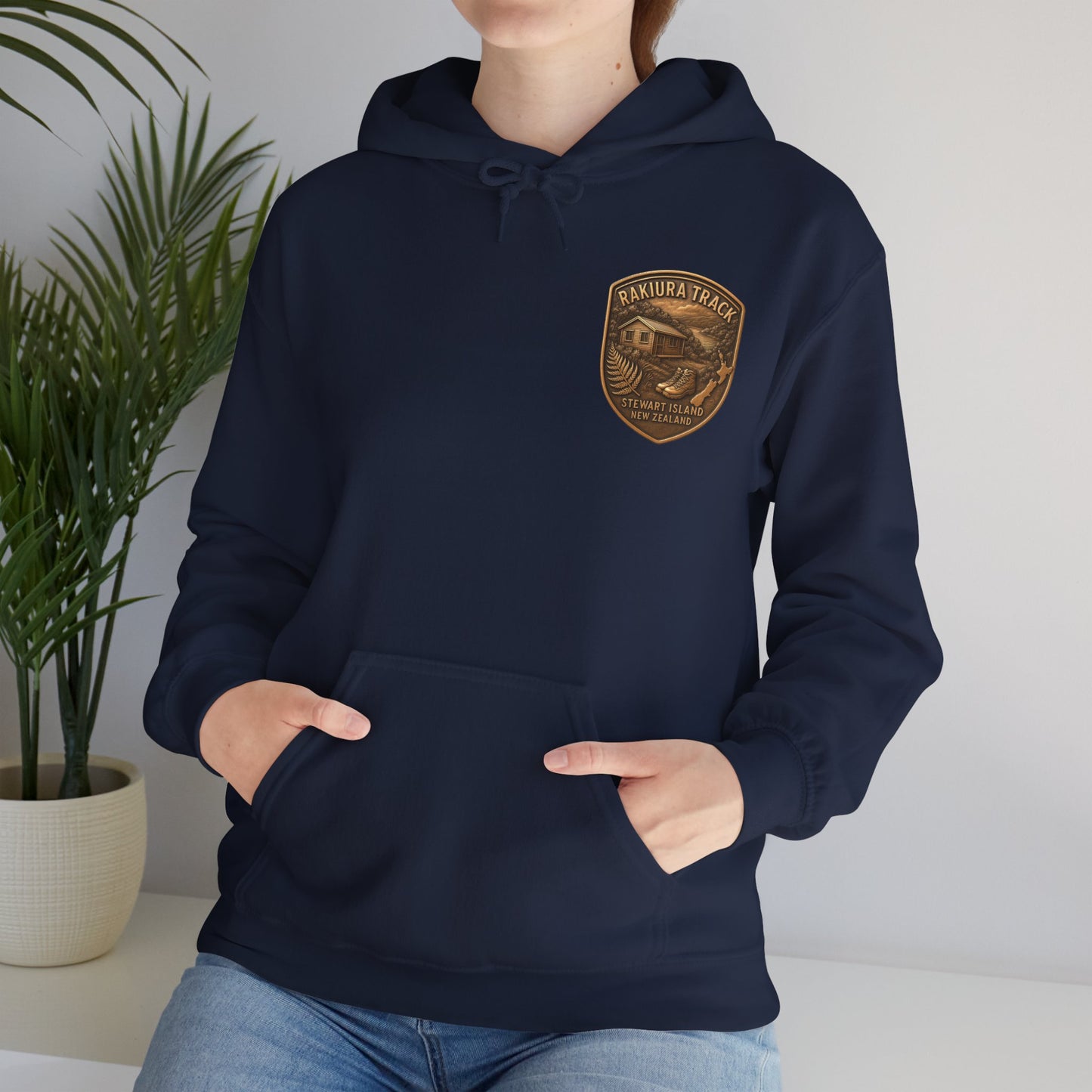 Person wearing a navy blue Rakiura Track hoodie with a gold emblem, standing indoors next to a plant.
