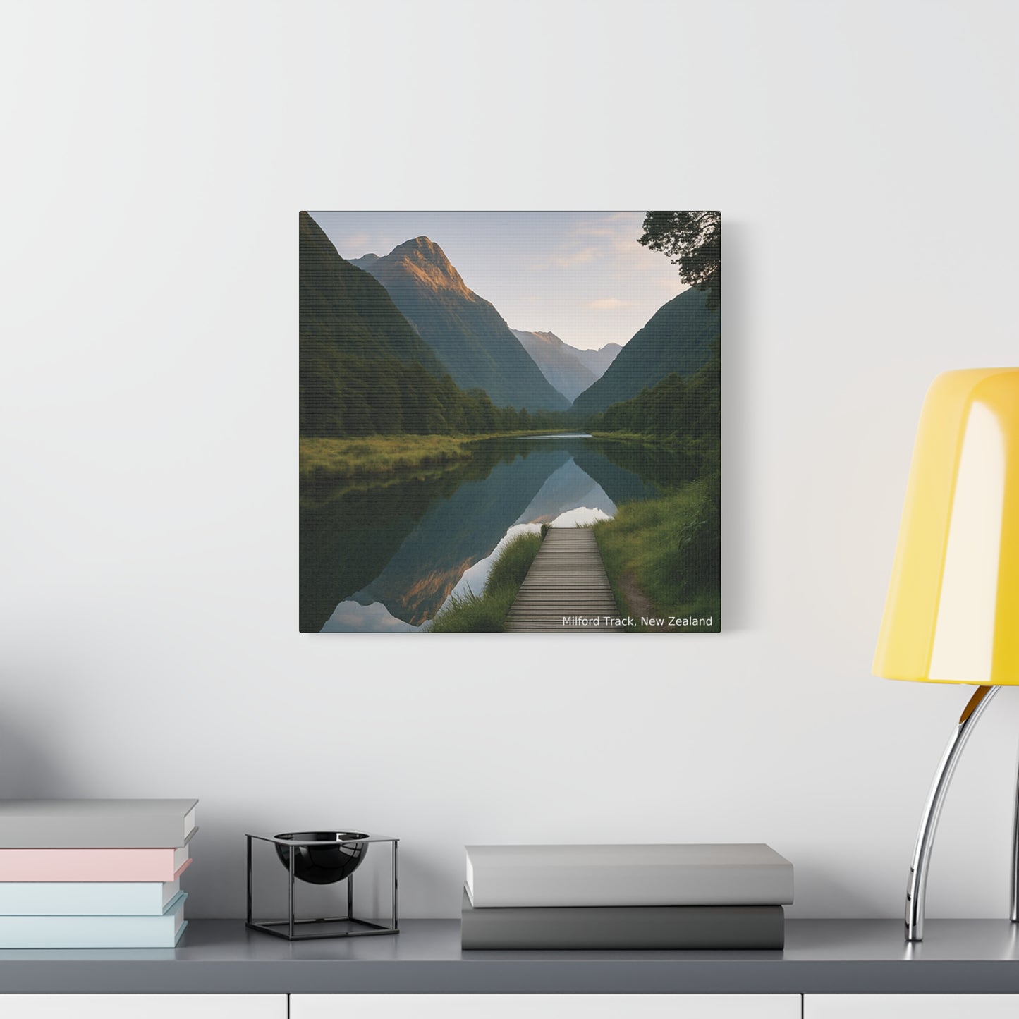 Framed landscape print of a mountainous valley with a river on a white wall above a shelf.