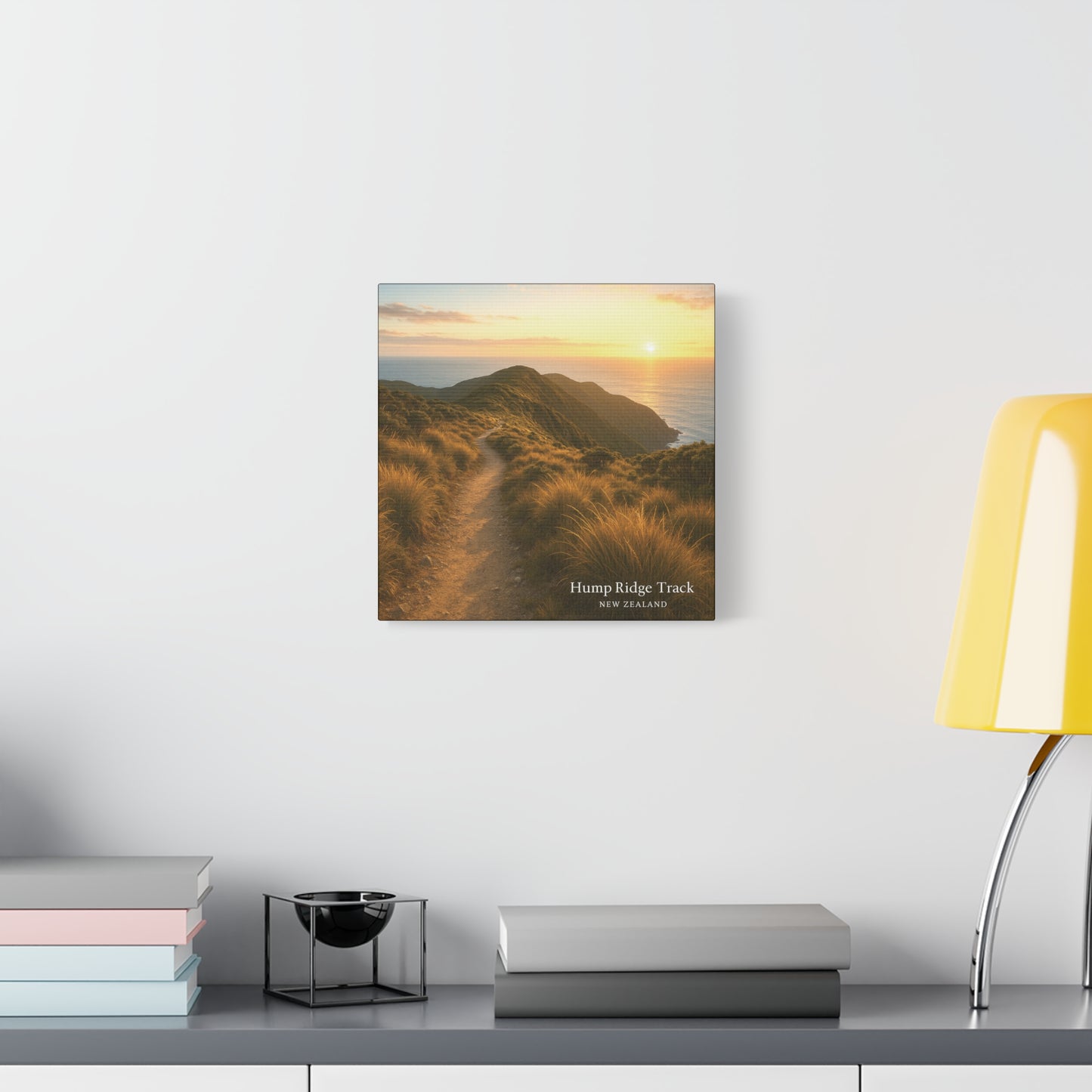 Hump Ridge Track Framed artwork of a sunset over a coastal landscape on a wall above a desk with books and a lamp.